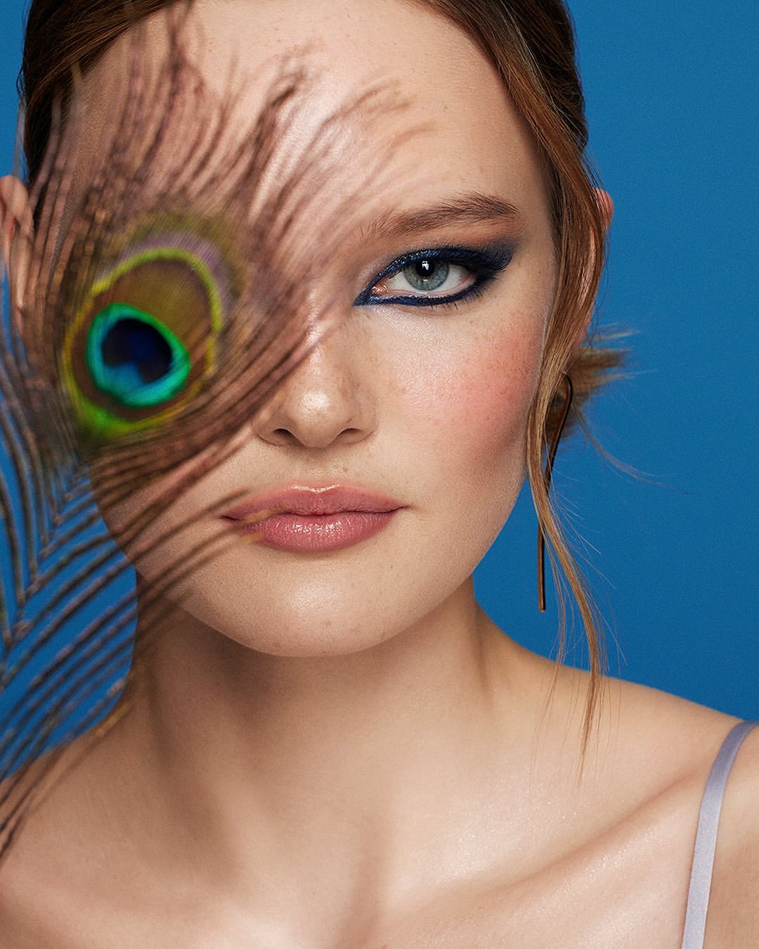 Close-up of a woman's face with blue eye makeup and a peacock feather partially covering her face against a blue background.