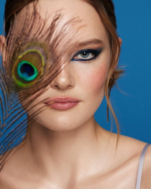 Close-up of a woman's face with blue eye makeup and a peacock feather partially covering her face against a blue background.