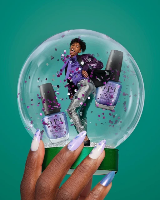 A person in a purple sequin jacket inside a snow globe, surrounded by nail polish and confetti against a teal background.