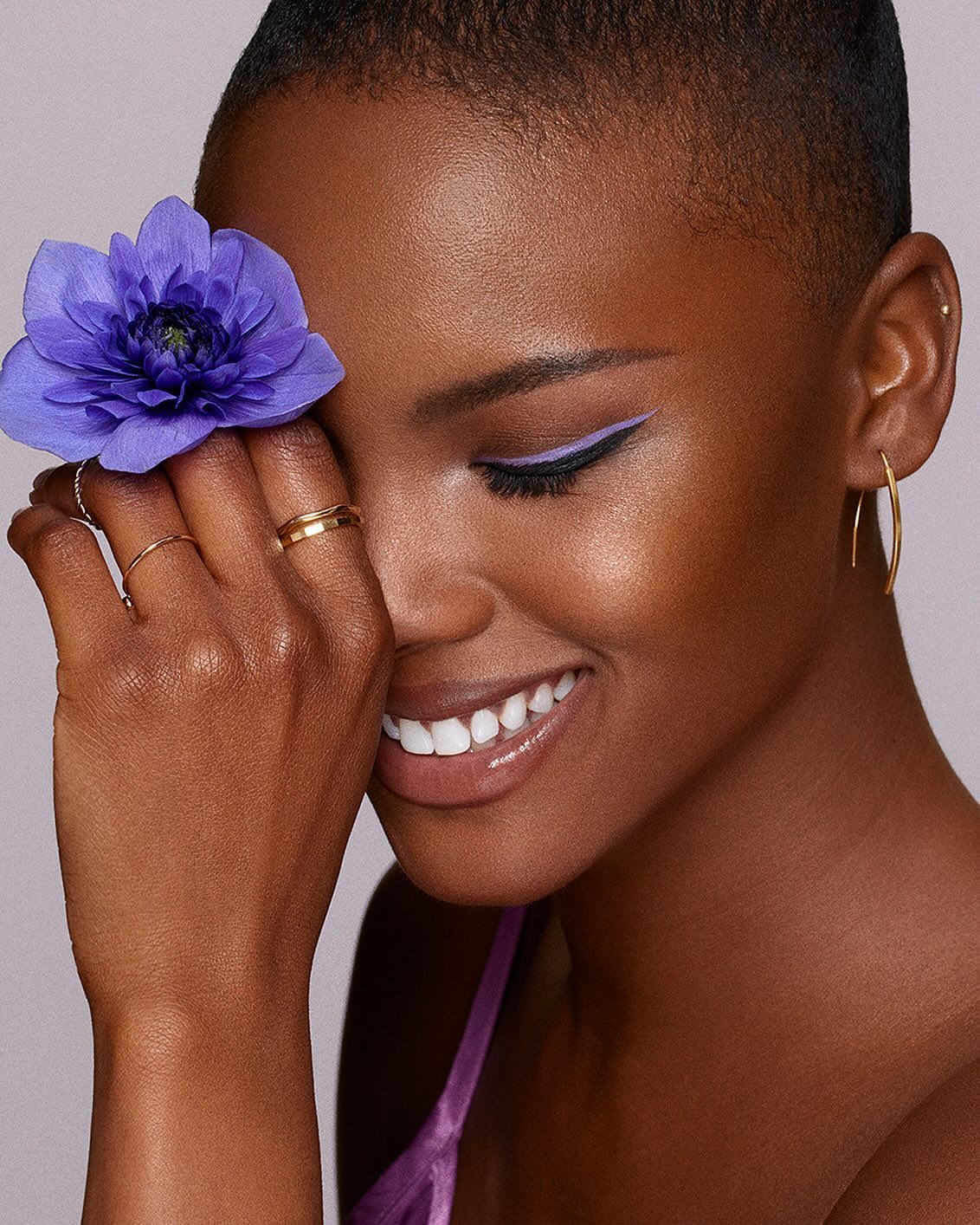 Close-up of a smiling woman with fair skin and a buzzcut, holding a purple flower to her face, featuring lilac eyeliner and gold jewelry.