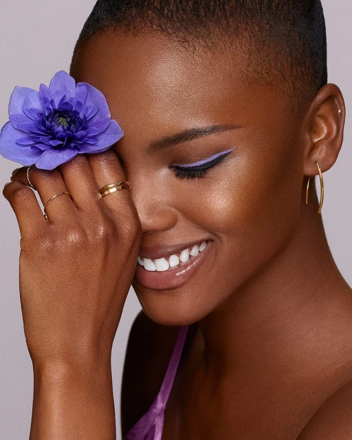 Close-up of a smiling woman with fair skin and a buzzcut, holding a purple flower to her face, featuring lilac eyeliner and gold jewelry.