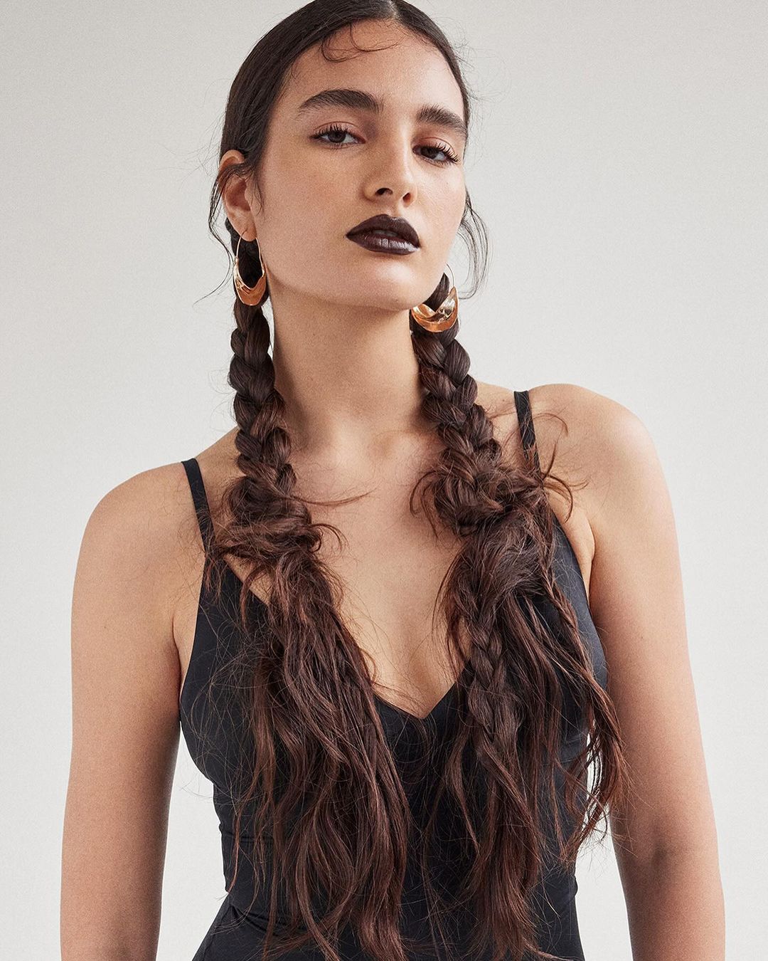 Portrait of a young adult woman with dark lipstick, wearing gold hoop earrings, and her long brown hair styled in pigtail braids.