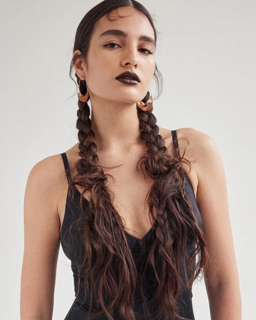Portrait of a young adult woman with dark lipstick, wearing gold hoop earrings, and her long brown hair styled in pigtail braids.
