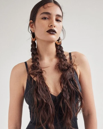 Portrait of a young adult woman with dark lipstick, wearing gold hoop earrings, and her long brown hair styled in pigtail braids.