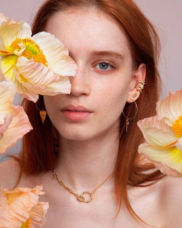 Close-up portrait of a red-haired woman framed by large pale peach poppy flowers, wearing gold jewelry.