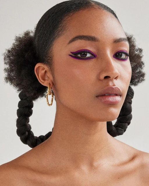Close-up of a young adult female with purple eyeliner, gold earrings, and braided pigtails against a neutral background.