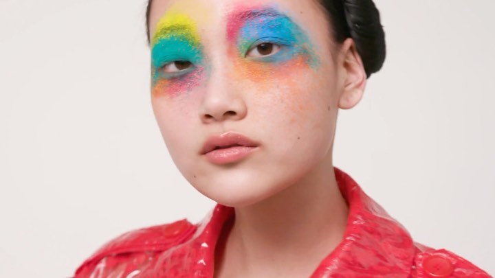 Close-up of a model wearing colorful makeup: yellow, pink, and blue eyeshadow in a beauty shot against a white background.