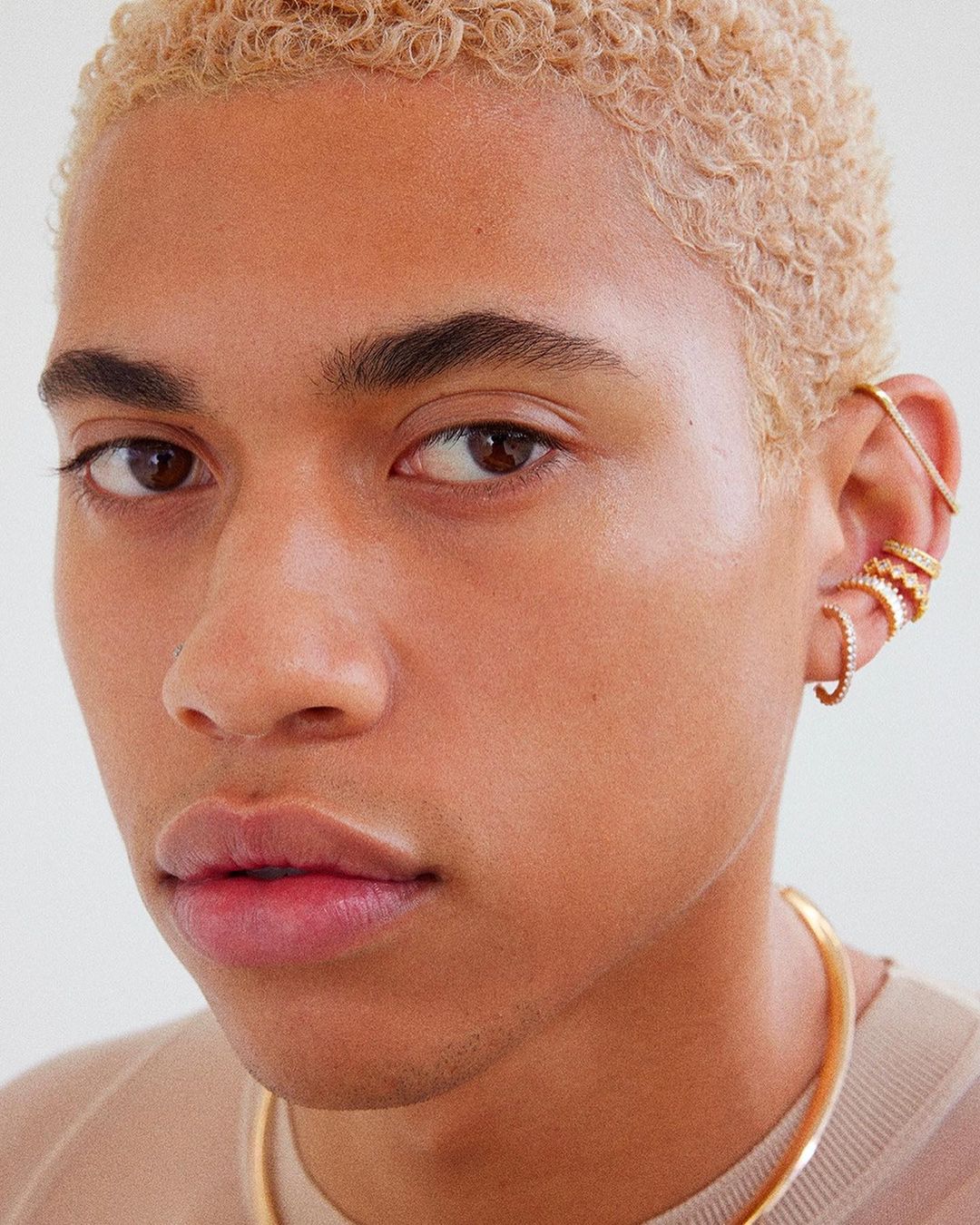 Close-up portrait of a young adult with short blonde hair and multiple gold earrings, set against a clean, bright backdrop.