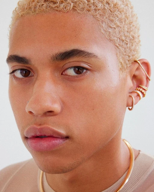 Close-up portrait of a young adult with short blonde hair and multiple gold earrings, set against a clean, bright backdrop.