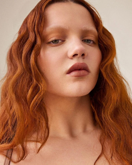 Close-up portrait of an adult woman with auburn wavy hair, neutral makeup, and fair skin against a neutral background, looking at the camera.