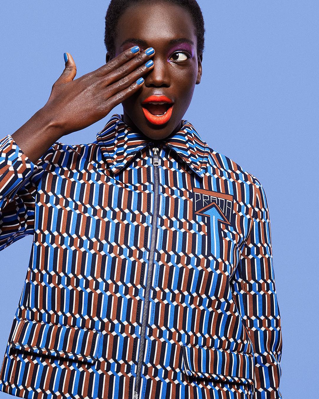 Fashion shot of an adult woman with short hair wearing a patterned Prada jacket against a light blue backdrop, covering one eye with her hand.