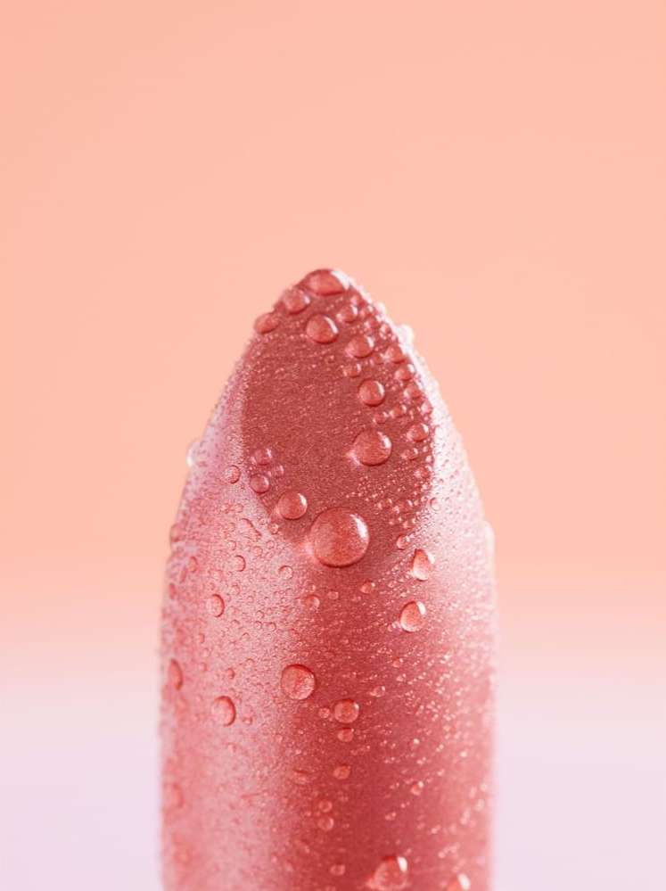 Close-up of a pink lipstick with water droplets on the surface. Beauty shot with a peach-colored background for cosmetic advertising.