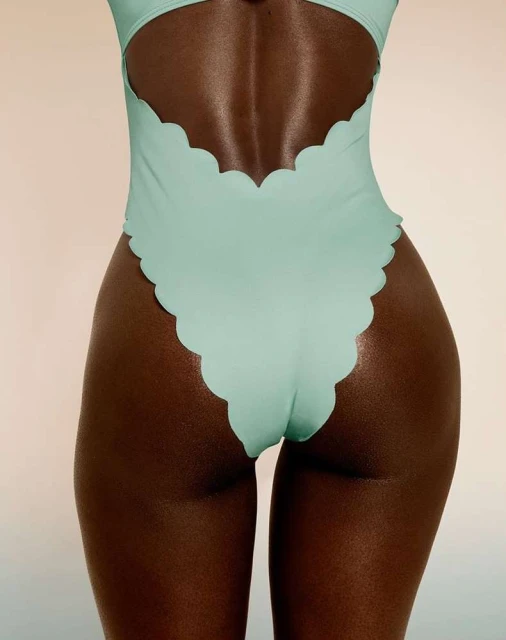 Close-up shot of a tan-skinned adult woman wearing a mint-green scalloped one-piece swimsuit against a light peach backdrop in portrait orientation.