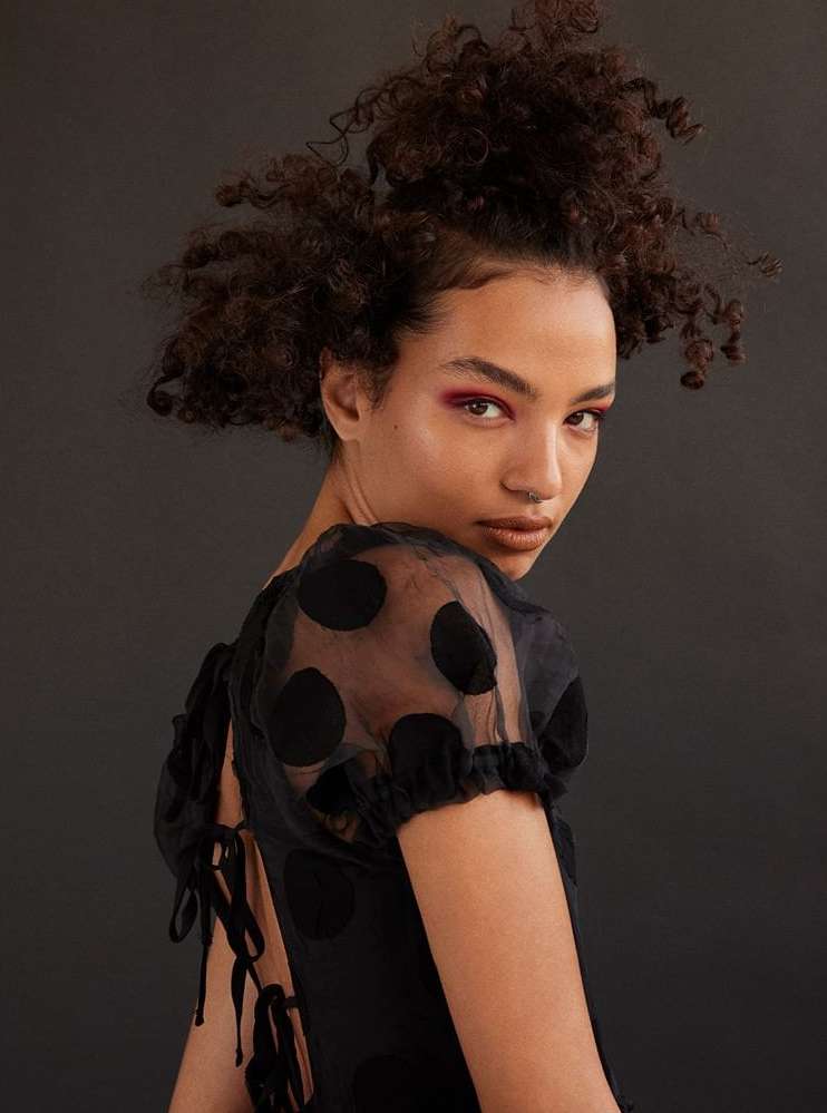 Woman with curly hair poses in a black polka dot dress against a dark background in a beauty shot.