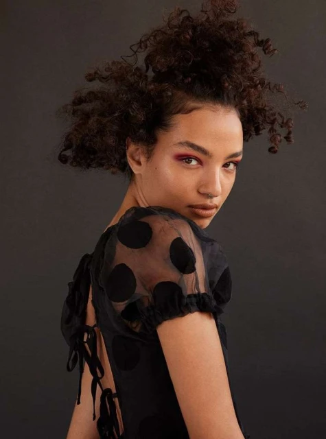 Woman with curly hair poses in a black polka dot dress against a dark background in a beauty shot.
