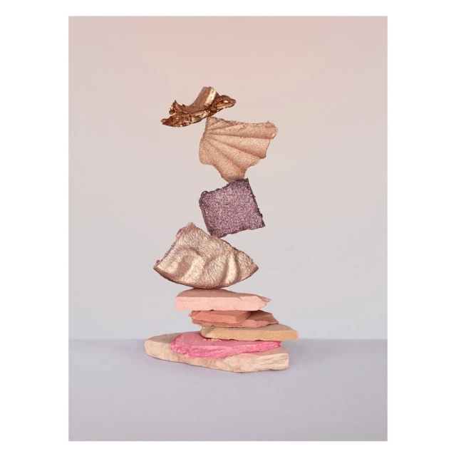 Creative still life of stacked, textured cosmetic swatches, featuring gold, pink, and purple tones against a pale background.