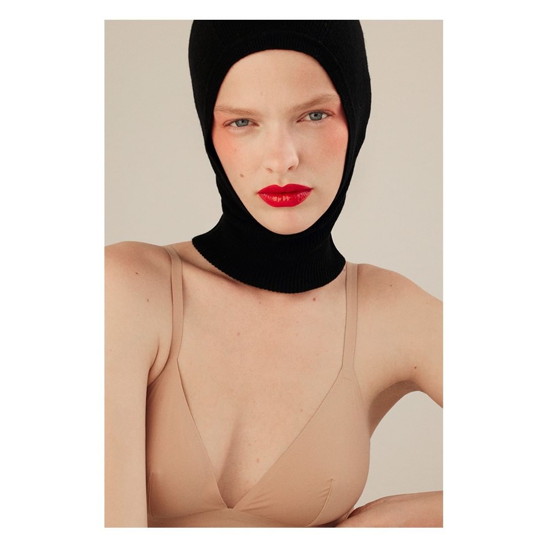 Close-up studio portrait of a female model with fair skin wearing a black knit balaclava and a beige bra against a neutral backdrop.