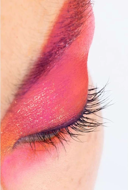 Close-up of a human eye with bright pink, orange, and gold eyeshadow, and long dark eyelashes against a stark white background.
