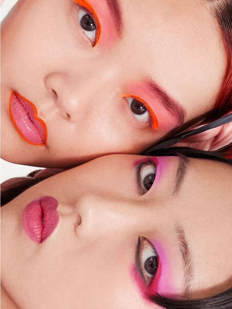 Close-up beauty shot features two Asian women with vibrant neon makeup: coral eyeshadow and lip liner, bright pink eyeshadow and lipstick.