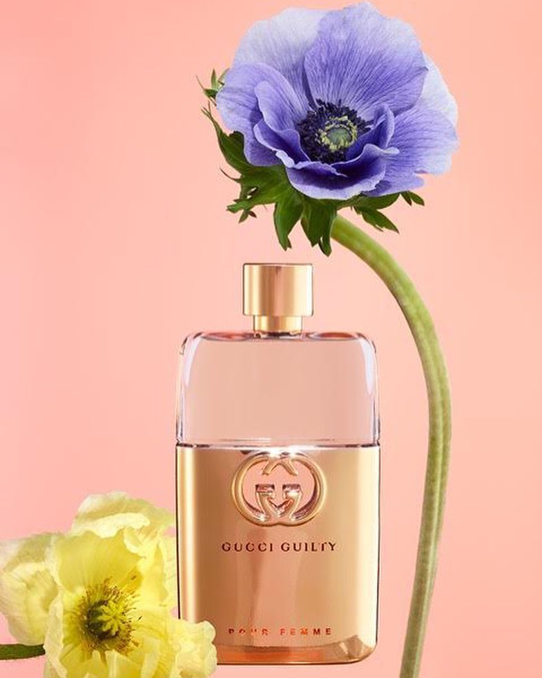 Gucci Guilty Pour Femme perfume bottle stands out with gold accents, complemented by purple and yellow flowers against a peach backdrop.