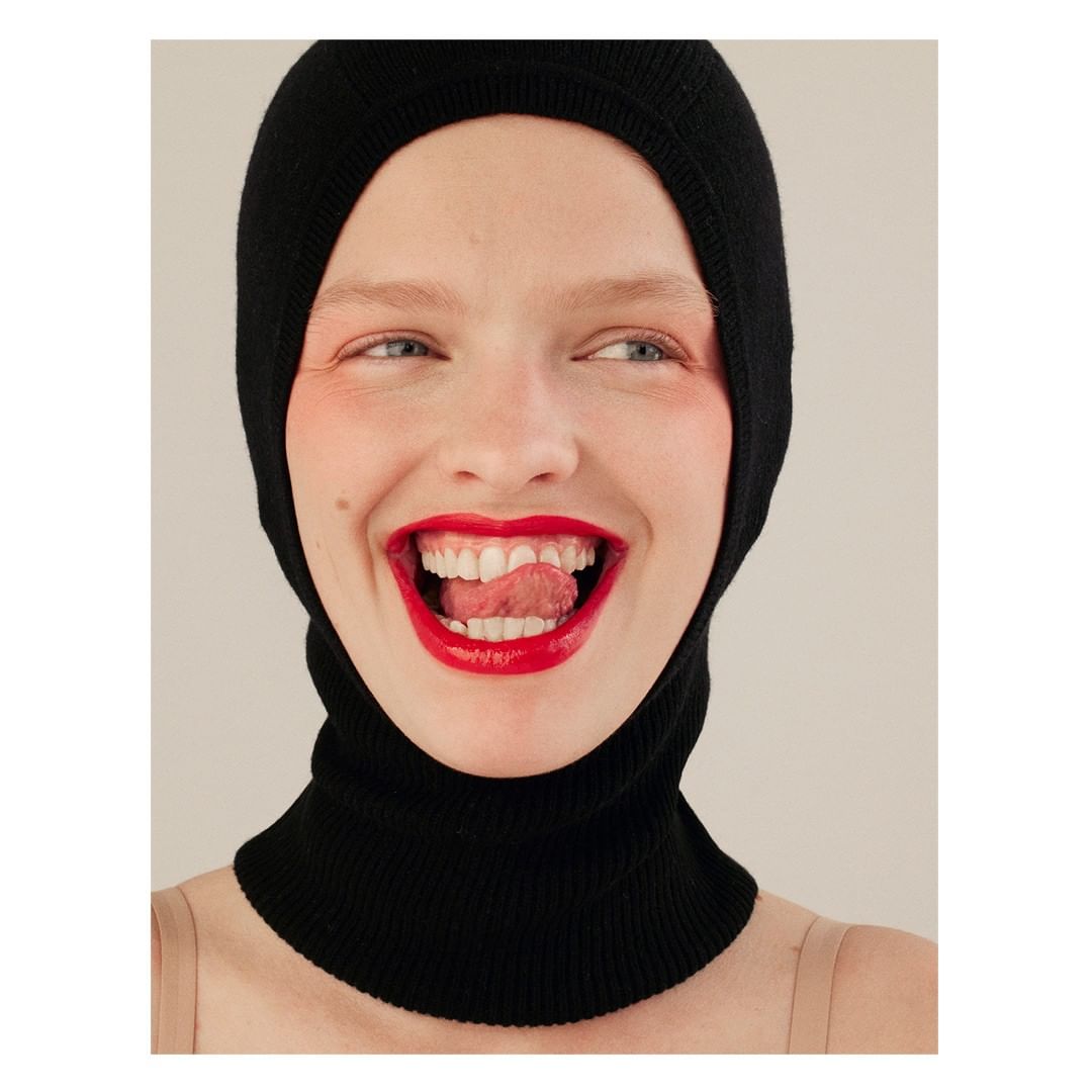 Close-up portrait of a smiling woman wearing a black knit balaclava and bright red lipstick, sticking out her tongue in a studio setting.