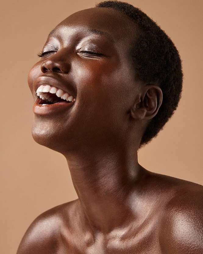 Close-up portrait of a dark-skinned Black woman with a shaved head laughing with her eyes closed in a beige photo studio setting.