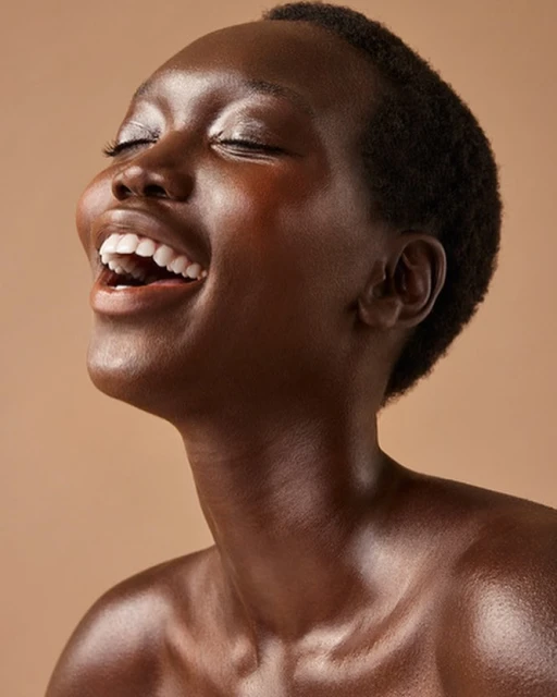 Close-up portrait of a dark-skinned Black woman with a shaved head laughing with her eyes closed in a beige photo studio setting.