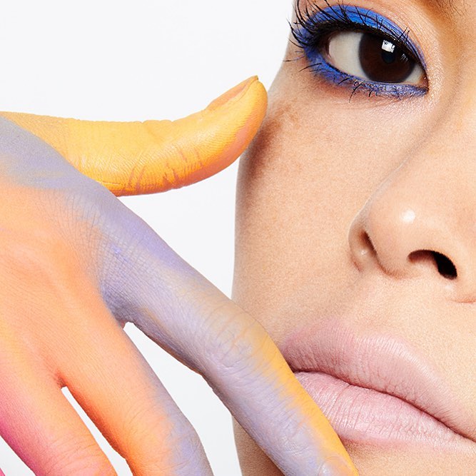 Close-up portrait of a person with blue eye makeup and colorful painted hand in a white background, emphasizing beauty and makeup.