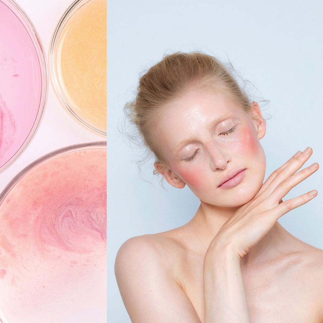 Fair-skinned woman with red blush poses near petri dishes filled with pink and orange liquids on a soft, light blue backdrop.