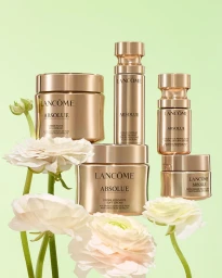 Group of Lancome Absolue skincare bottles and jars in rose gold packaging arranged with white ranunculus flowers on a pastel green background