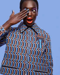 Black woman with blue nail polish covering her eye while wearing a geometric patterned jacket against a blue background