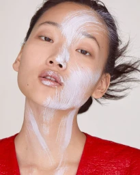 Close-up portrait of a woman with white paint smudged across her face and neck, wearing a red top against a neutral background.
