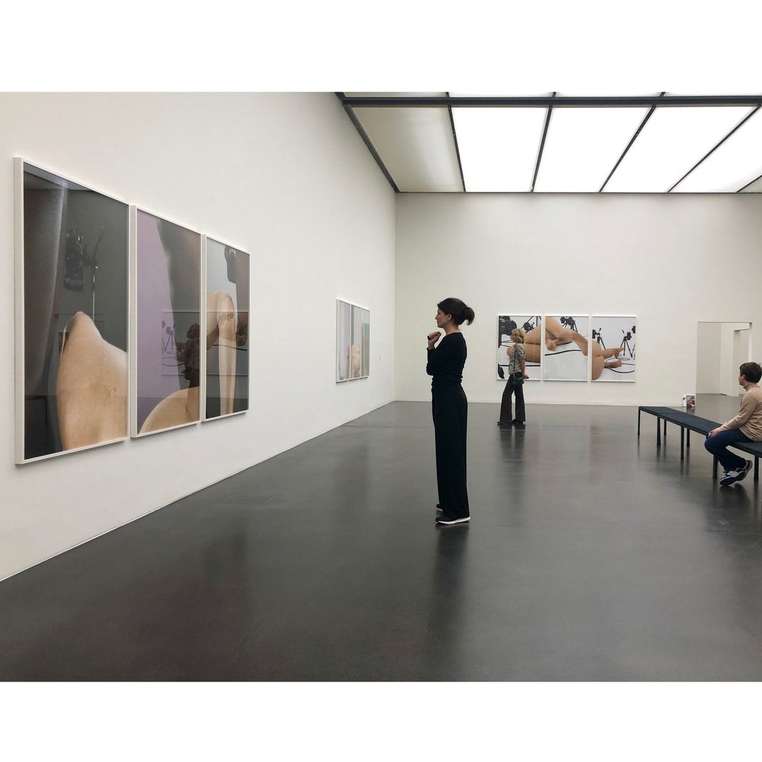 Woman views large-scale nude photographs in a museum gallery with white walls, black floors, and bright overhead lighting.