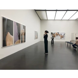 Woman in black top and trousers standing in a white art gallery viewing large framed photography triptychs on the wall.