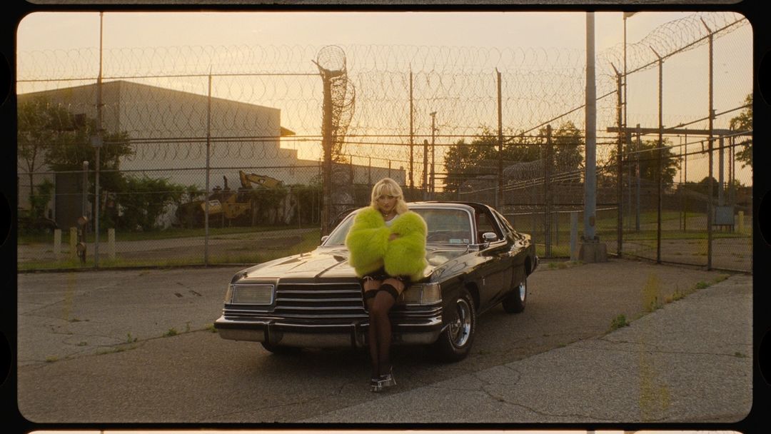 Blonde woman poses on a black car in a vibrant green faux fur coat against a chain-link fence in an outdoor setting at sunset.
