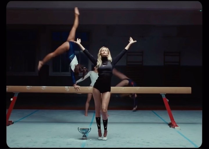 Adult woman in black sportswear stands in front of gymnasts doing balancing beam exercises in an indoor gymnasium setting.
