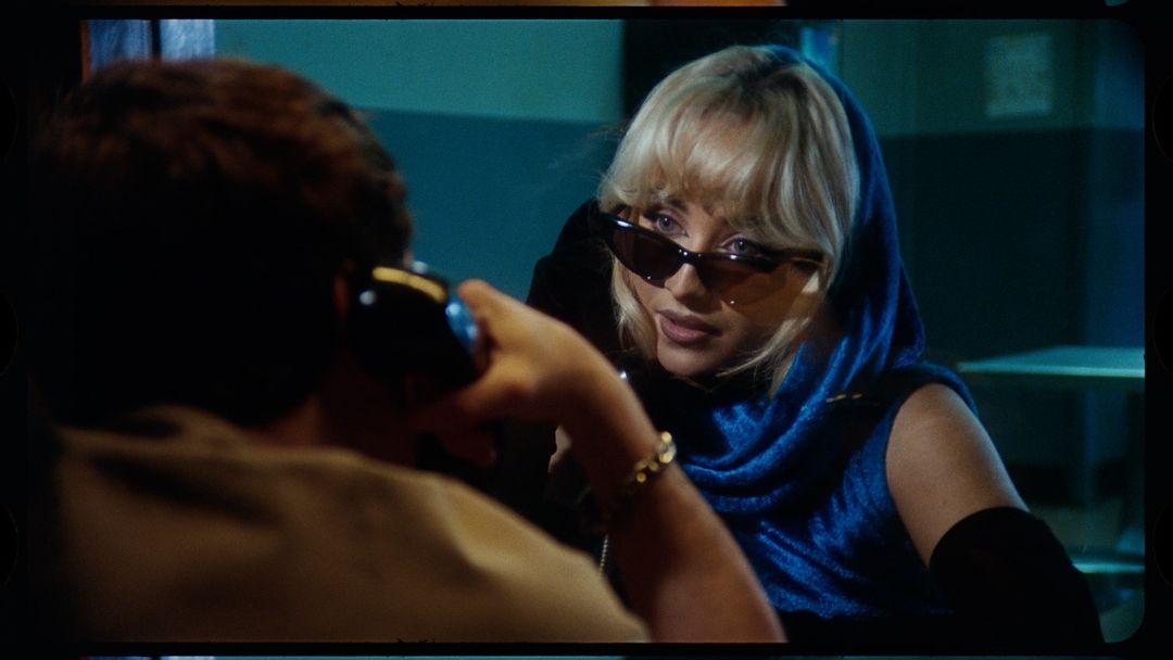 Woman in sunglasses with scarf speaking on a vintage phone, and another person is on the opposite side, indoor setting, eye-level view.