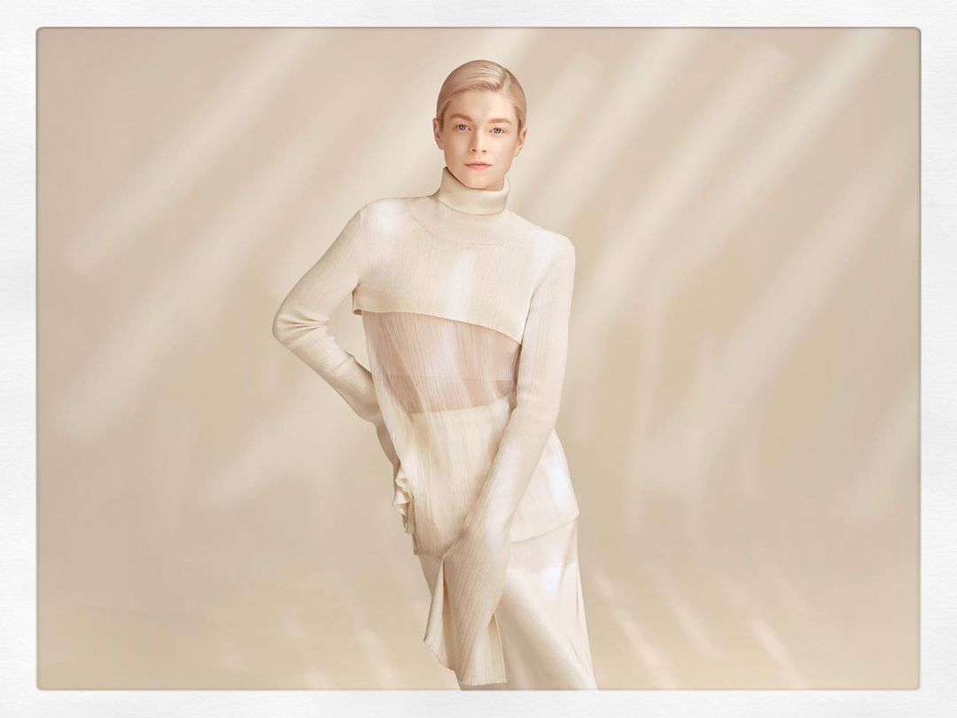 A poised adult female fashion model with blonde hair stands indoors against a neutral backdrop, showcasing an elegant cream-colored outfit in soft lighting.