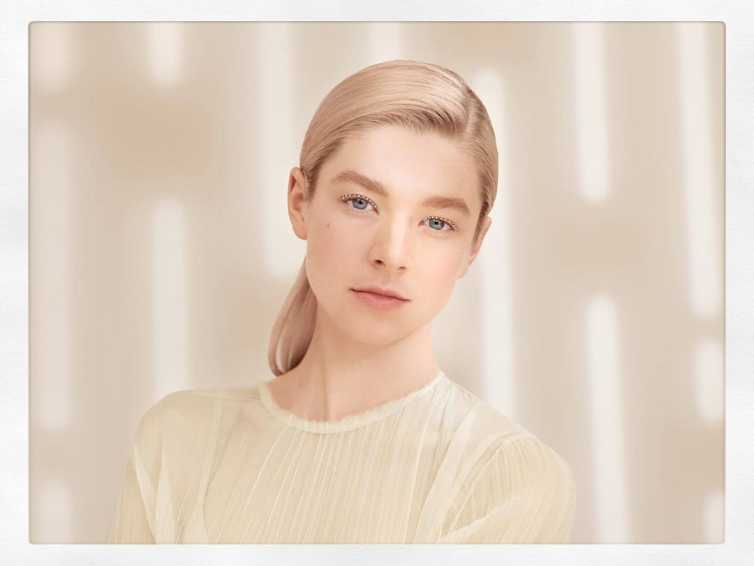 Close-up of a fair-skinned young woman with blonde hair slicked back and wearing a light, sheer top against a neutral, patterned background.
