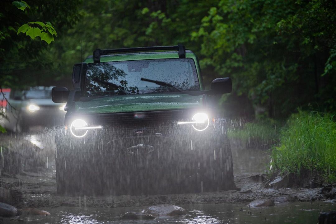 Green Ford Bronco SUV driving through a muddy puddle in a wooded area with its headlights on during a rainy, overcast day.