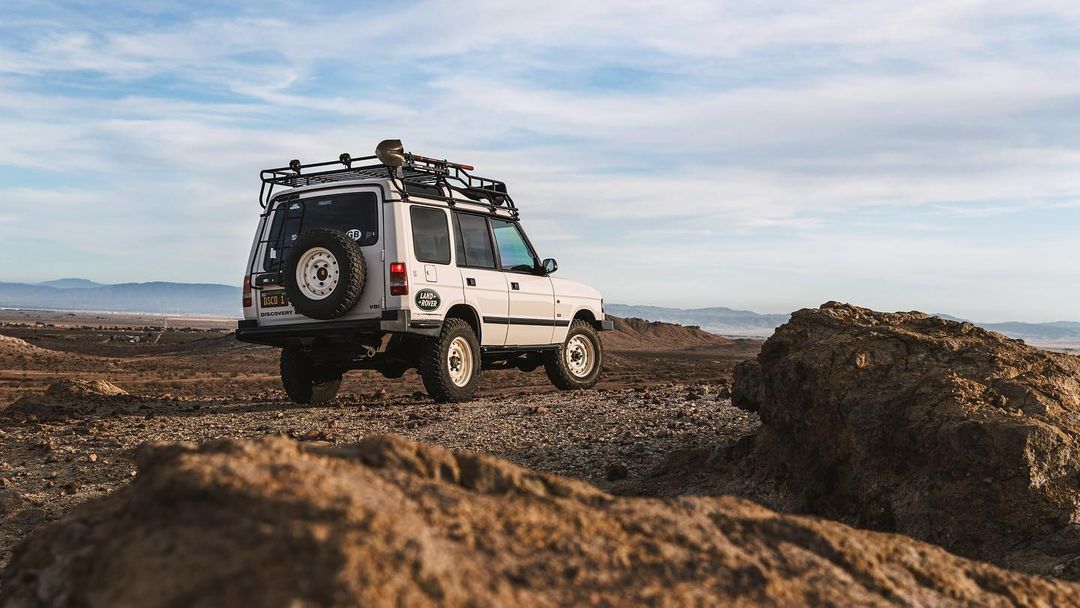White Land Rover Discovery parked on a rocky, desert landscape with blue sky and light cloud cover, offering an outdoor adventure aesthetic.