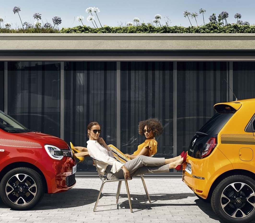 Two women lounge between a red and yellow Renault Twingo car in an outdoor setting with modern building facade and green roof.