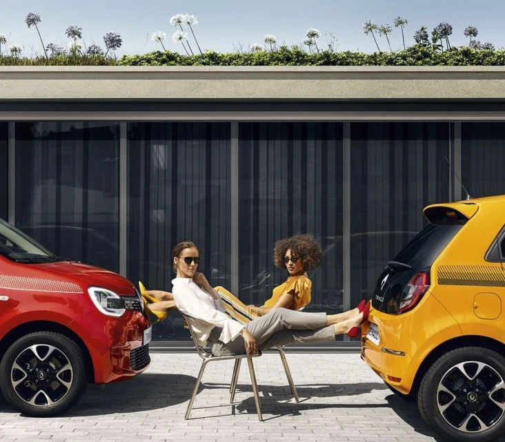 Two women lounge between a red and yellow Renault Twingo car in an outdoor setting with modern building facade and green roof.