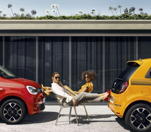 Two women lounge between a red and yellow Renault Twingo car in an outdoor setting with modern building facade and green roof.