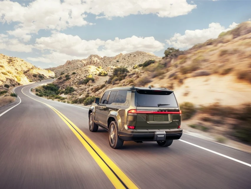 Rear view of a green SUV driving on a desert highway with mountains and a blue sky with clouds in the background, motion blur present.