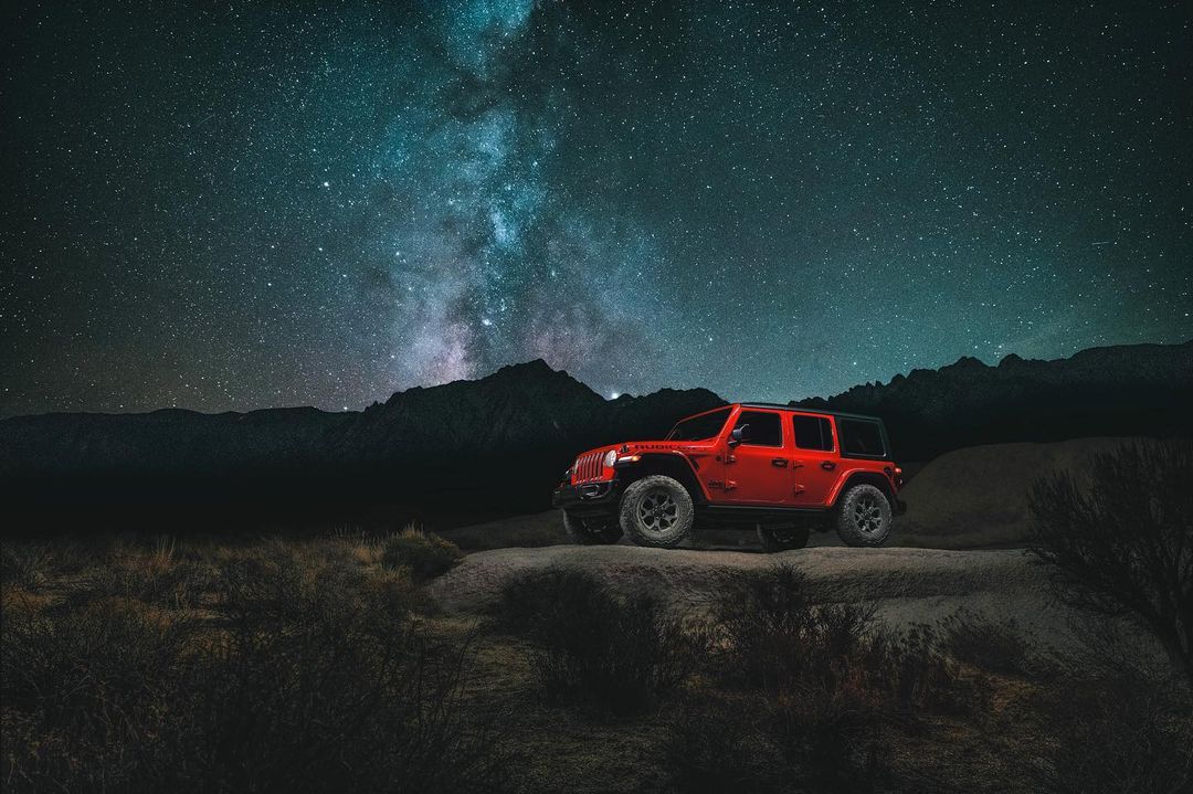 An orange Jeep Wrangler stands on a rocky outcrop under a night sky full of stars and the Milky Way, mountains in the background.
