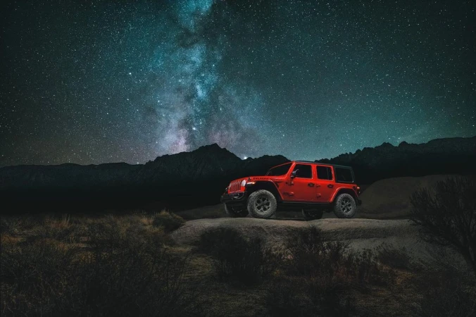 An orange Jeep Wrangler stands on a rocky outcrop under a night sky full of stars and the Milky Way, mountains in the background.