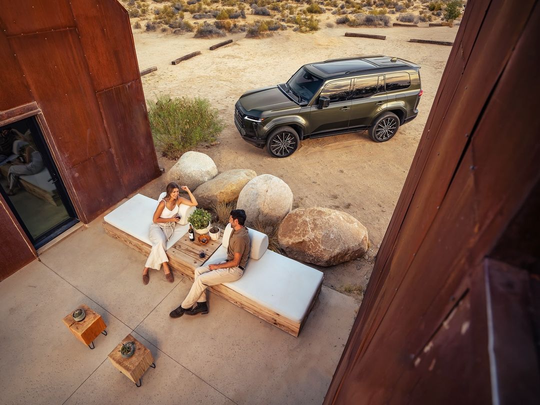 Olive-green SUV parked near desert dwelling with a couple relaxing on a white daybed on the patio; arid desert setting.