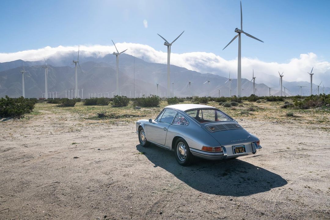 Silver Porsche parked on a desert plain with many wind turbines and a mountain range in the distance under a partly cloudy sky.