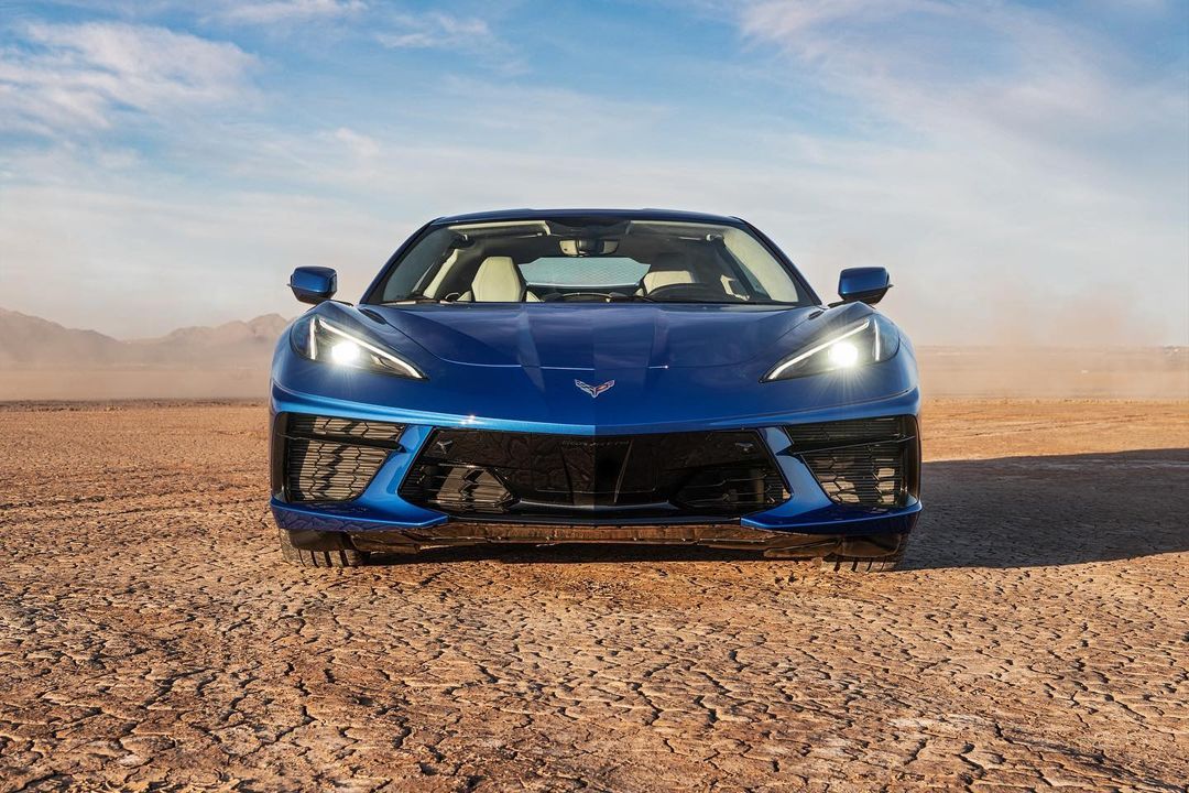 Blue Corvette parked on cracked dry land with a desert and mountains in the background under a blue sky with scattered clouds.